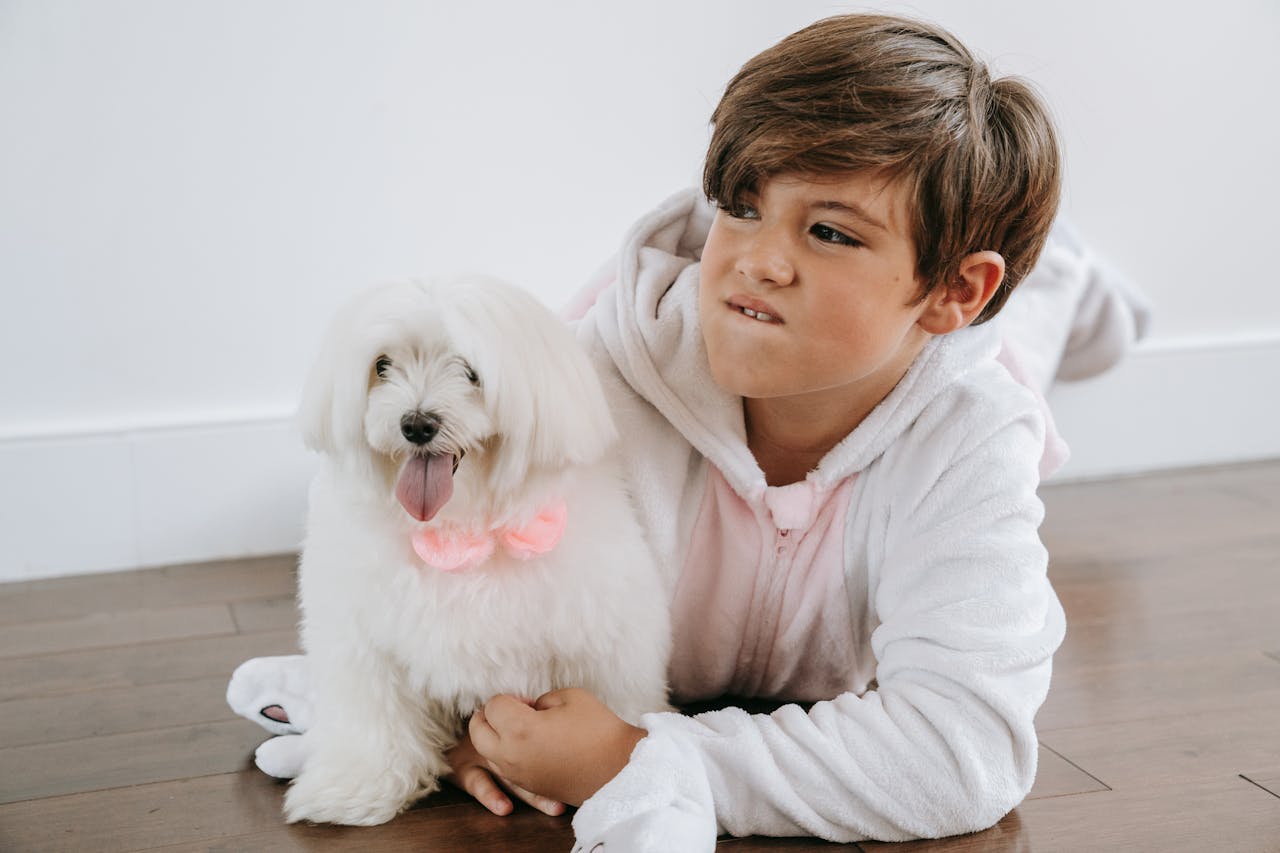 A child dressed in a cozy costume lies on the floor with a fluffy Maltese dog.