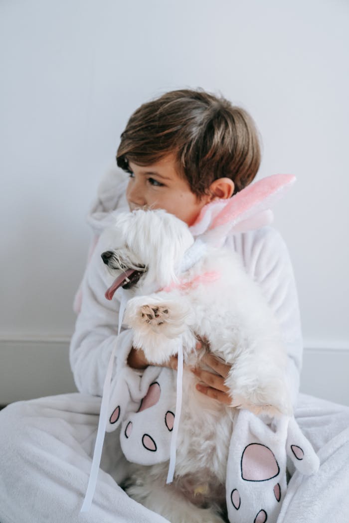 Young boy in a bunny onesie holding a fluffy Maltese dog indoors.
